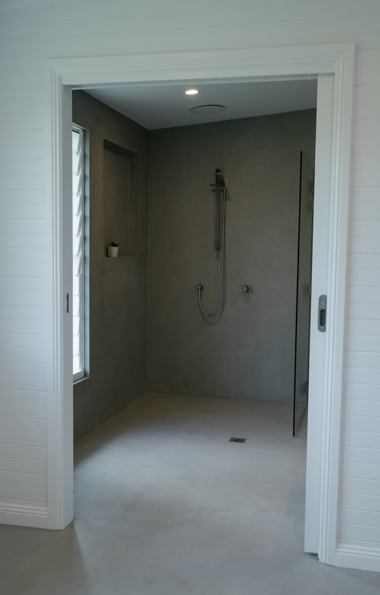 View through a doorway into a microcement shower room with a louvre window